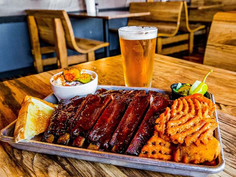 A plated BBQ ribs dinner with sweet potato fries, cornbread, and a cold beer.