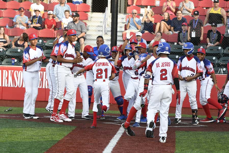 Dominican baseball team celebrates at home plate during a game with enthusiastic players and fans.