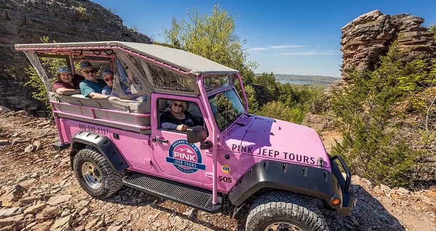 A bright pink Jeep tour vehicle filled with passengers stops among rocky cliffs and trees.